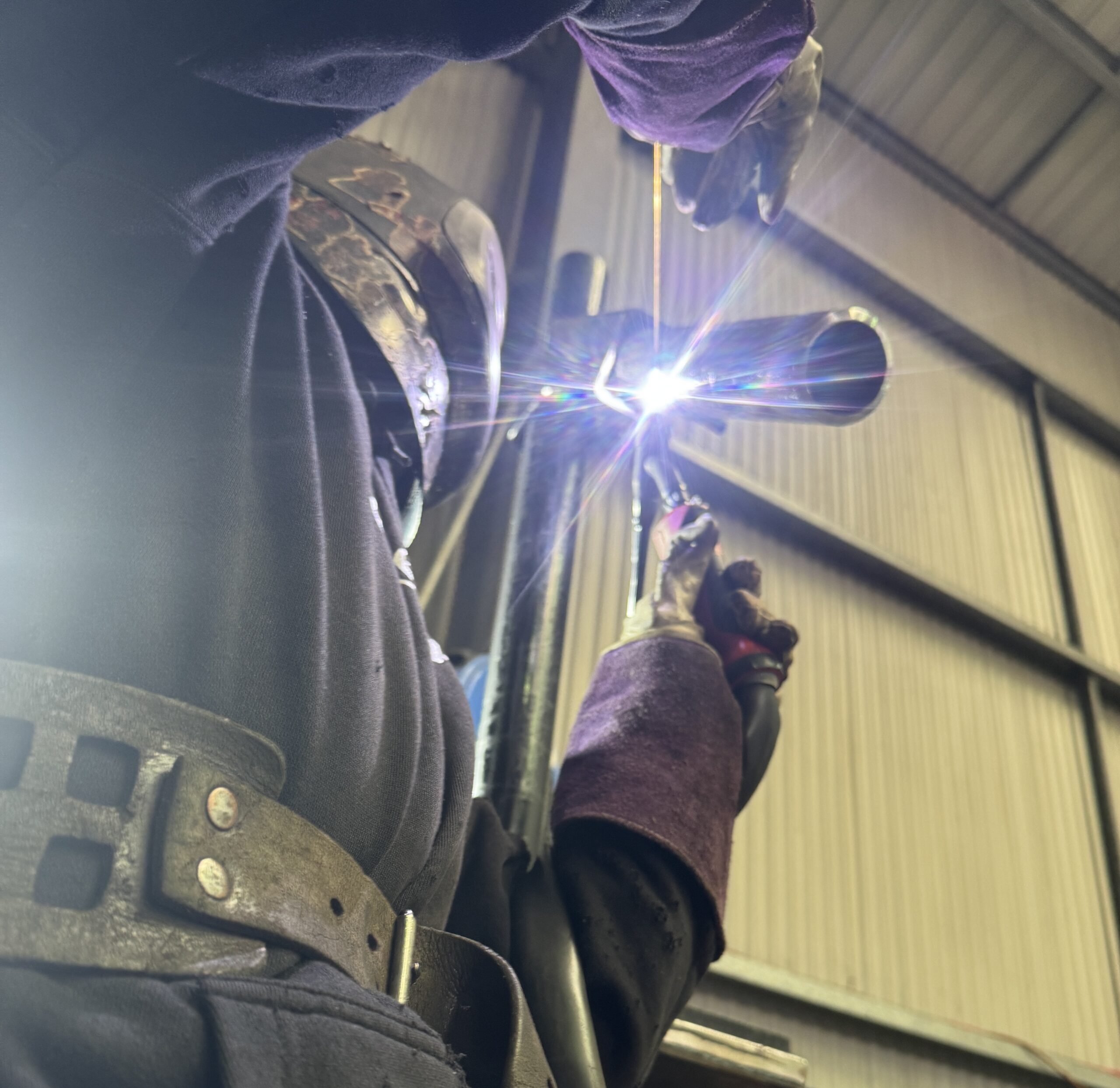 A person wearing protective gear and a welding helmet is welding a metal pipe, with bright sparks visible. The photo is taken from a low angle inside an industrial metal building.