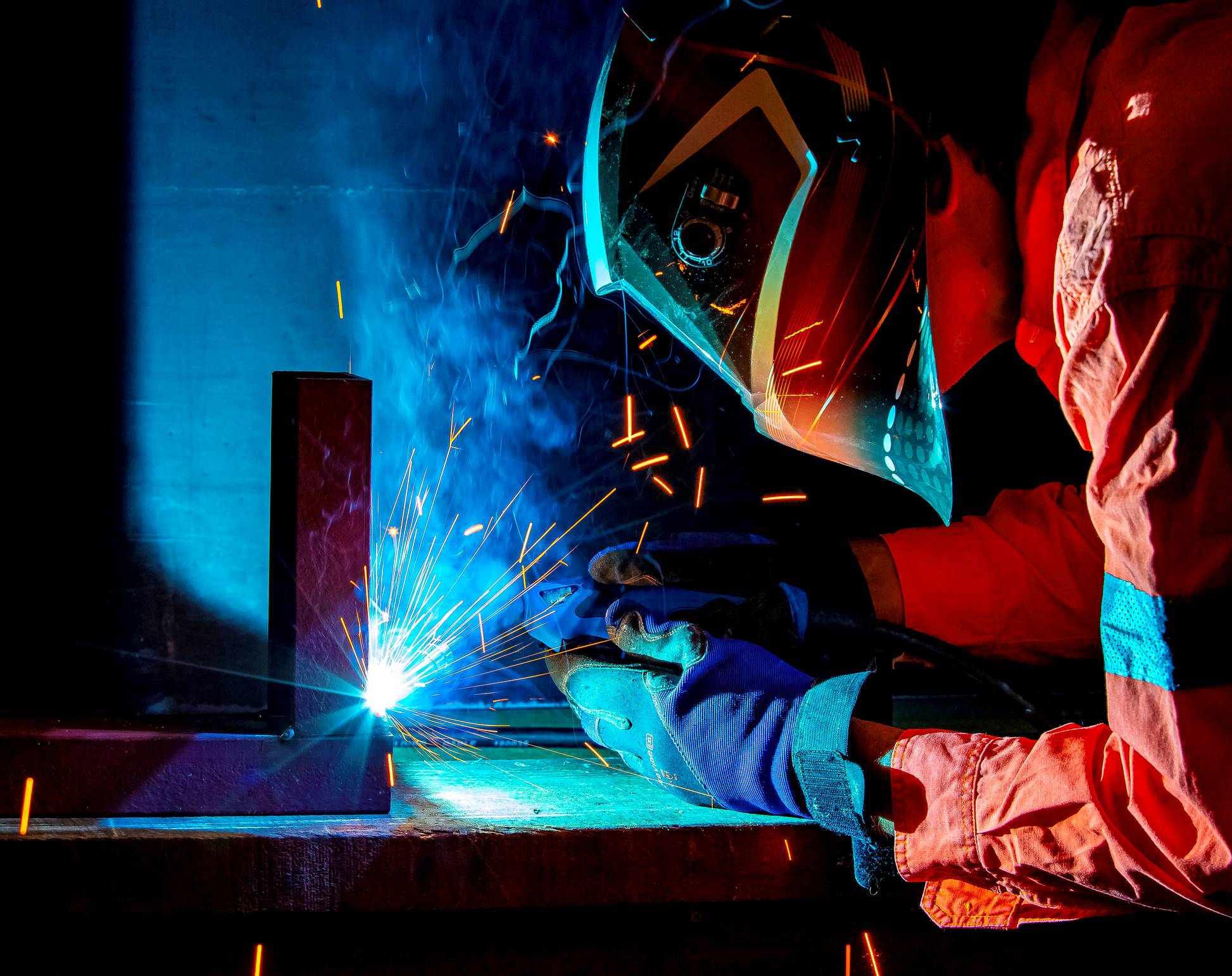 A person wearing protective gear and gloves is welding metal, with bright blue sparks and light emitted from the welding process in a dark workshop.