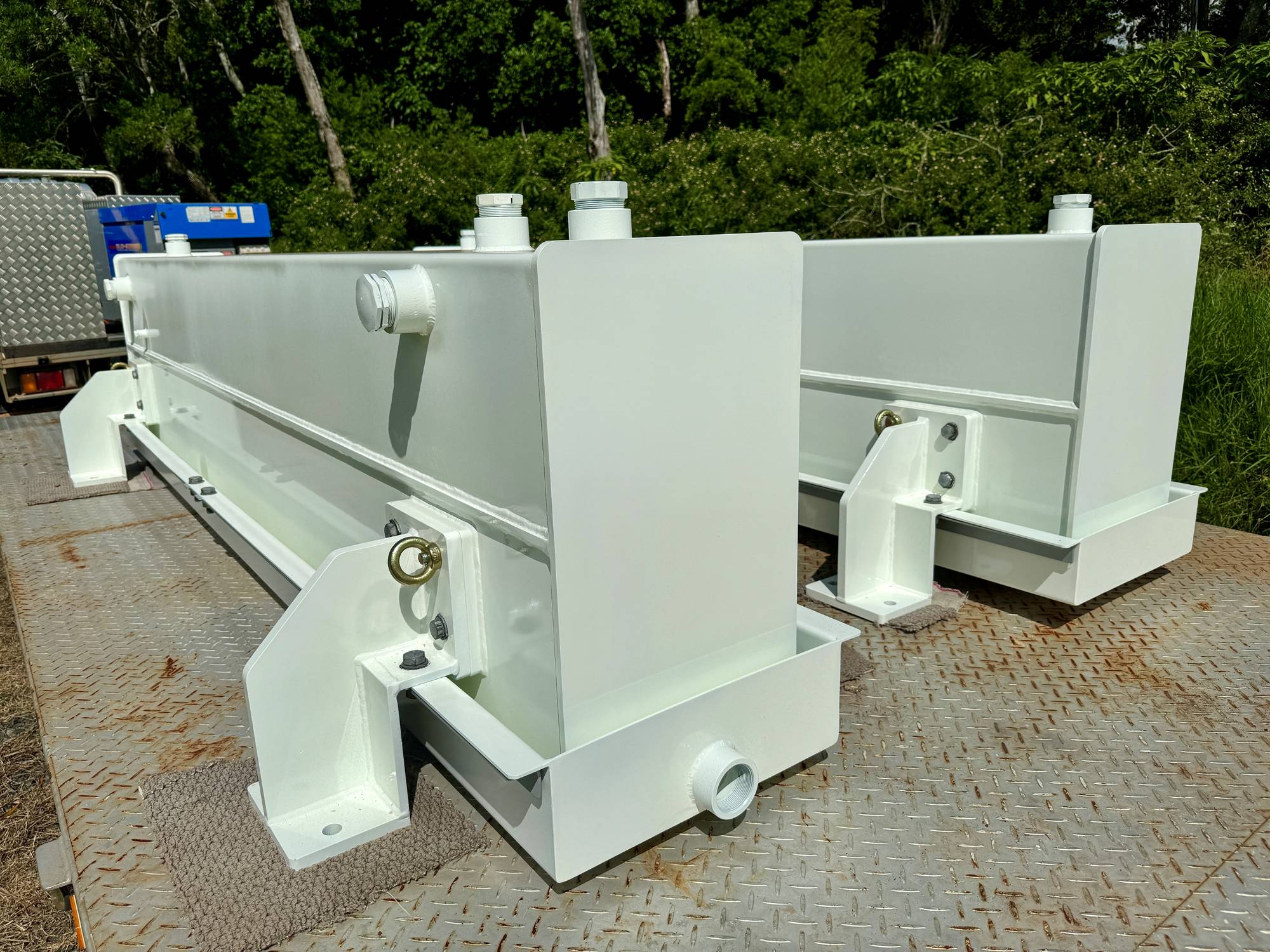 Two large, white rectangular industrial tanks with pipes and mounting brackets sit on a metal platform outdoors, surrounded by greenery and trees in the background.