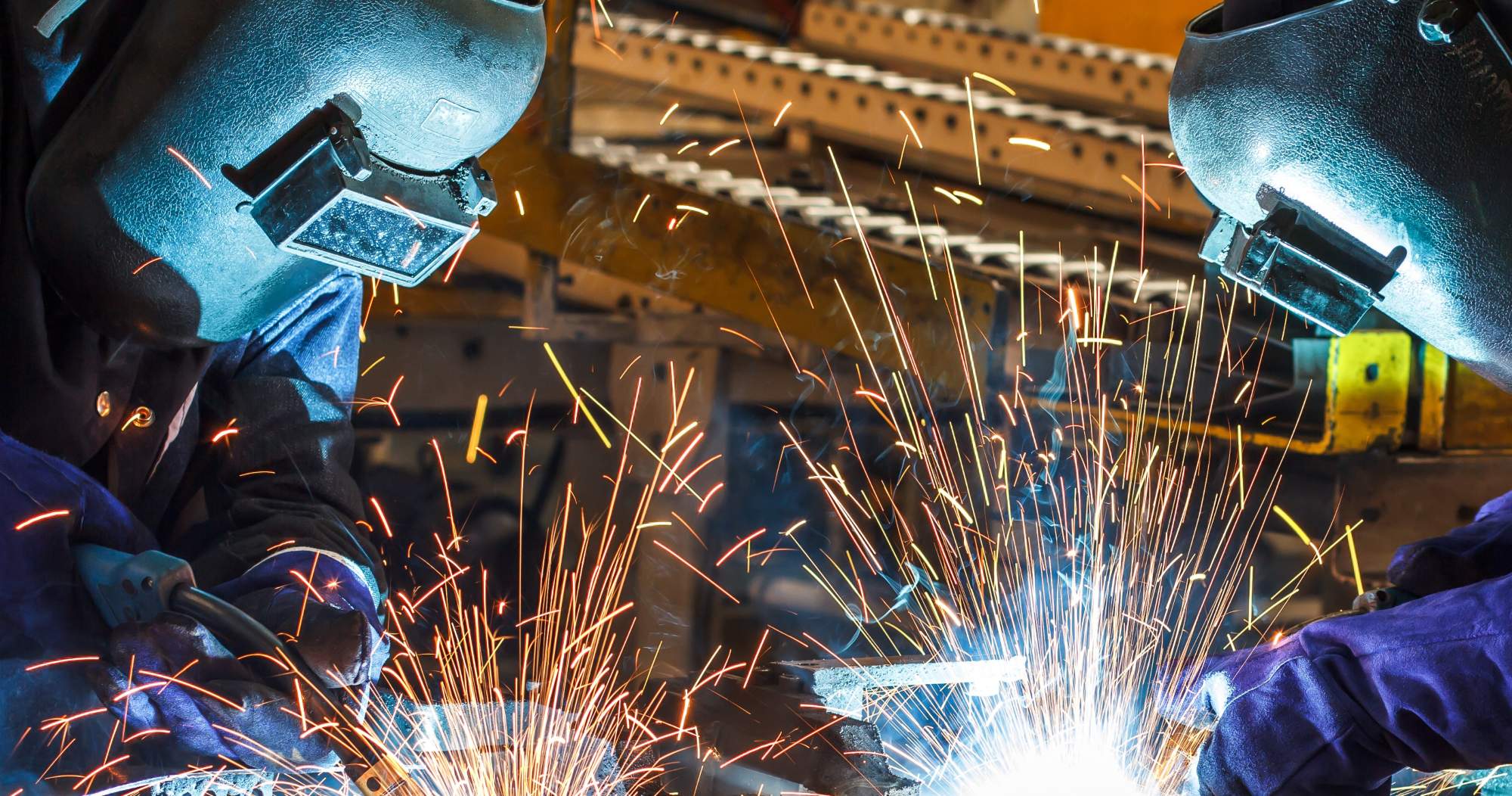 Two people wearing protective welding helmets and gloves are working with metal, producing bright sparks and light in an industrial setting.