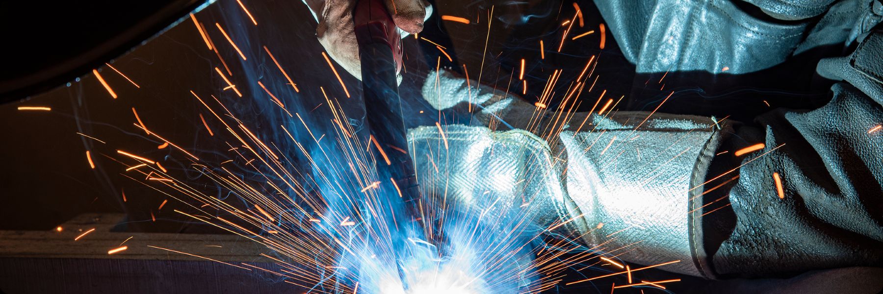 A person wearing protective gloves and gear is welding metal, producing bright blue light and orange sparks.