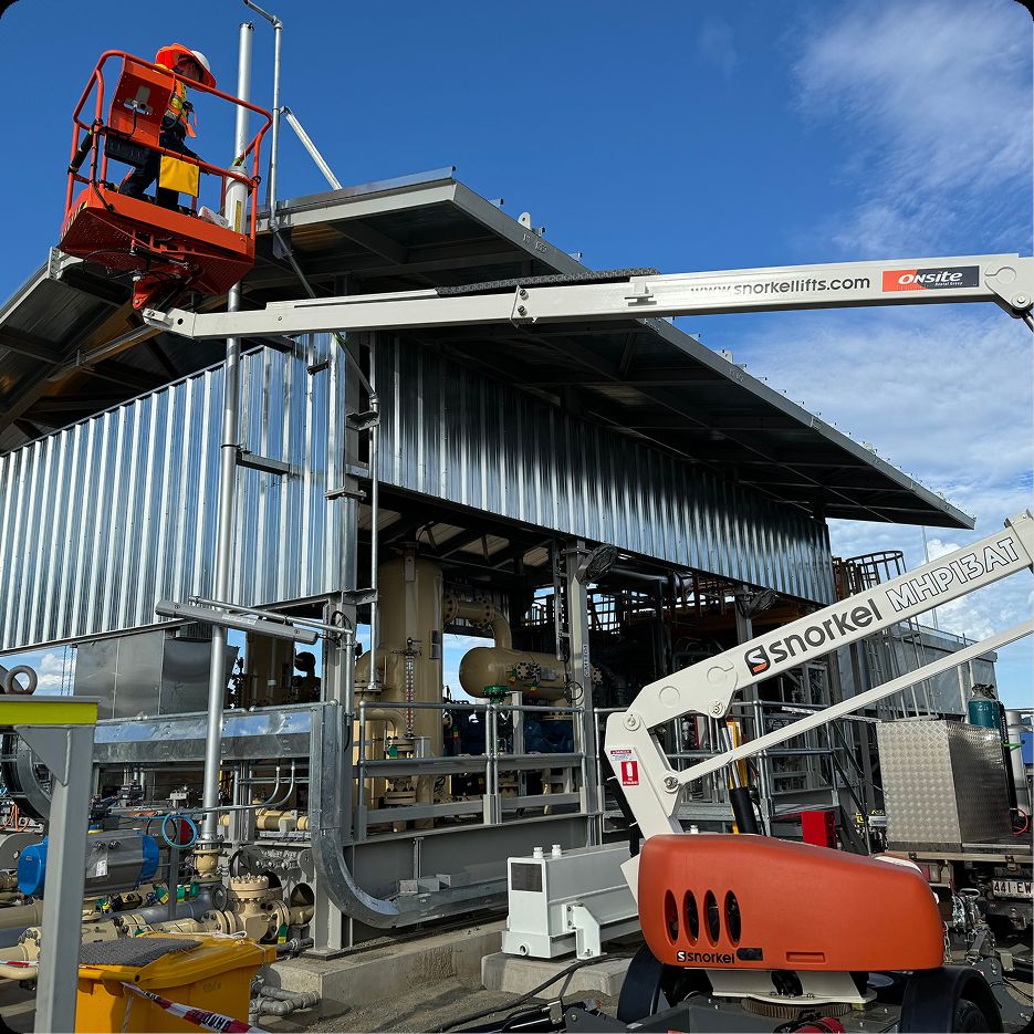 A worker in safety gear stands on a raised orange lift beside a large metal industrial building, with machinery and pipes nearby under a blue sky. A white Snorkel lift is also positioned in front of the structure.