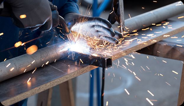 A person wearing protective gloves uses a welding torch to join two metal pipes, producing bright sparks and a blue glow during the welding process.