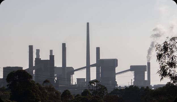 The image shows the silhouette of an industrial plant with tall chimneys and structures, some emitting smoke, set against a hazy sky. Trees and power lines are visible in the foreground.