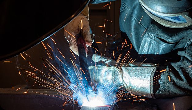 A person wearing protective gear welds metal, producing bright blue and orange sparks in a workshop setting.