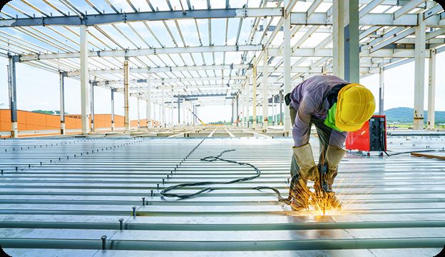 A construction worker wearing a yellow hard hat and protective gloves welds metal beams on the floor of a large, unfinished industrial building. Bright sparks are visible from the welding process.