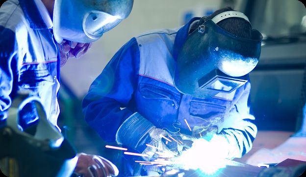 Two people wearing protective welding masks and gear are welding metal, with bright blue sparks and light visible from the welding process.